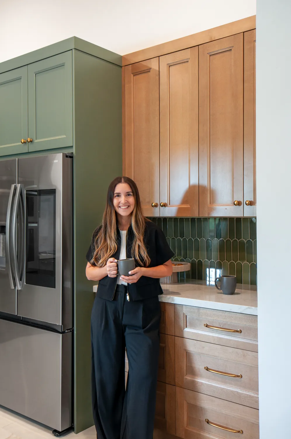 Camila Borges Chiang holding a coffee in a kitchen she designed, with sage green cabinetry